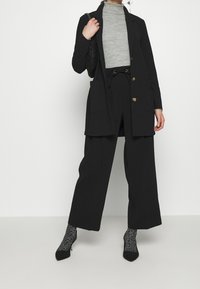 Černý elegantní blazer s dvěma předními kapsami, šedý rolák, černé kalhoty se širokými nohavicemi a černé lodičky s vzorovanými ponožkami.