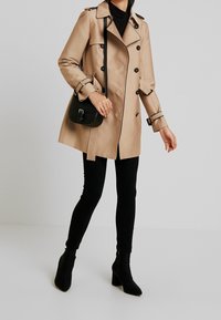 Beżowy płaszcz typu trencz z dwurzędowym zapięciem, czarnymi guzikami i paskiem w talii. Do tego dopasowane czarne spodnie i czarne botki na obcasie.