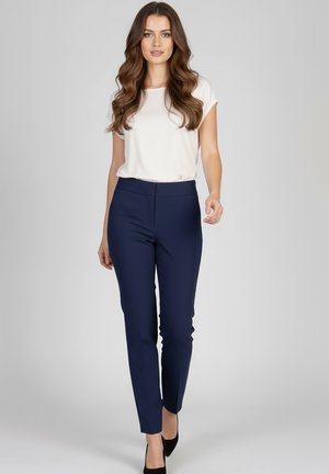 Kobieta idąca do przodu, ubrana w biały krótki rękaw, dopasowane granatowe spodnie i czarne szpilki na jednolitym szarym tle.