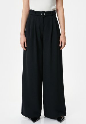 Schwarze Schlaghosen mit einem Gürtel, mit Falten, maßgeschneiderter Passform und leichtem Stoff. Abgerundet mit spitz zulaufenden schwarzen Schuhen.