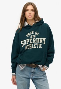 Sudadera con capucha en color verde azulado con la impresión "SUPERDRY ATHLETIC" en color blanco sucio, bolsillo frontal, llevada sobre una camiseta de rayas, combinada con jeans azul desgastado.