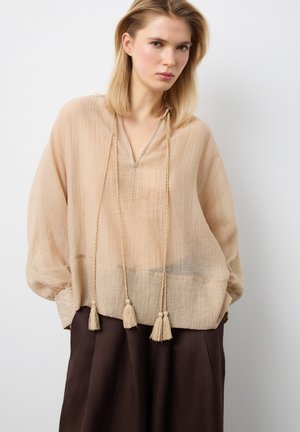 Mujer rubia con una blusa beige suelta con cordones de borlas y pantalones oscuros de pierna ancha, de pie frente a un fondo claro y liso.