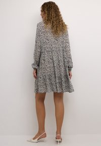 Robe à motifs floraux en tissu léger avec taille froncée, manches longues et ourlet volants. Présente des couleurs marine, crème et taupe ; accompagnée de talons blancs.