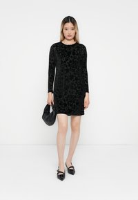 Donna che indossa un vestito lungo nero in velluto floreale con maniche lunghe, tiene una borsa nera e indossa delle scarpe nere a punta su uno sfondo bianco.