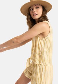 Top senza maniche a righe gialle e bianche abbinato a pantaloni corti coordinati. Tessuto leggero con vestibilità ampia e dettaglio del nodo frontale. Cappello a falda larga.