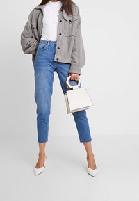 Šedá kostkovaná oversize bunda, bílý žebrovaný top, modré slim džíny, bílé lodičky na vysokém podpatku a malá bílá kabelka s dřevěným úchytem.