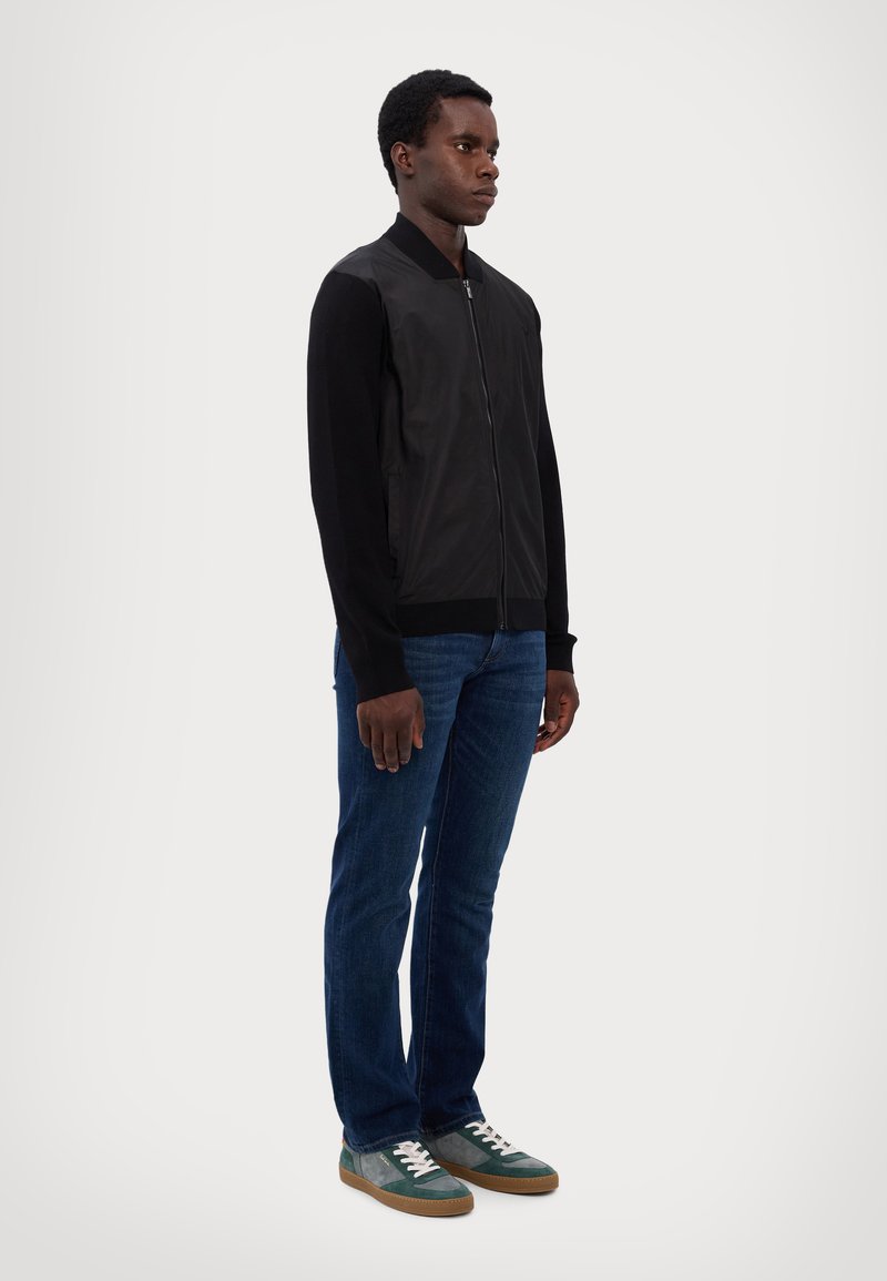 Black bomber jacket with leather front, knitted sleeves, zip closure. Worn with blue denim jeans and green sneakers with white laces.