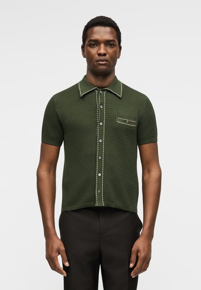 Άνδρας που φοράει σκούρο πράσινο πλεκτό πουκάμισο με κοντά μανίκια, λευκές ραφές, κουμπιά μπροστά και μια τσέπη στο στήθος με κουμπί.
