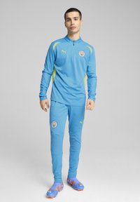 Gaiši zils sporta kostīms ar dzeltenām akcentēm, ar rāvējslēdzēja apkakli, logotipu uz krūtīm, pieguļošām biksēm un spilgti zilām sporta kurpēm ar oranžiem akcentiem.