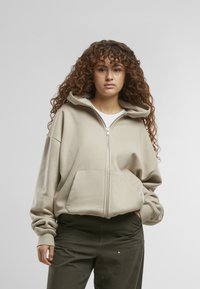 Beige zip-up hoodie met een grote capuchon, een losse pasvorm en voorkruiszakken. De stof lijkt zacht met manchetten aan de mouwen en een eenvoudig ontwerp.