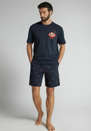 Man draagt een marineblauw T-shirt met een illustratie van een slapende hond en bijpassende marineblauwe korte broek met kleine rode hondenpatronen, staand op blote voeten.