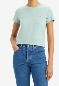 Světle mátově zelené bavlněné tričko s kulatým výstřihem a krátkými rukávy, v kombinaci s modrými džíny s vysokým pasem. Červené logo na hrudi.