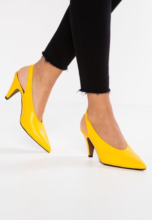 Żółte sandały na obcasie z odkrytą piętą, z czubkiem w szpic, błyszczącym wykończeniem i wąskim drewnianym obcasem. Noszone z czarnymi krótko przyciętymi dżinsami.