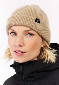 Tan ribbed beanie van gebreid materiaal, voorzien van een zwart label met witte tekst. Draagt bij een zwarte jas en toont een strakke pasvorm.