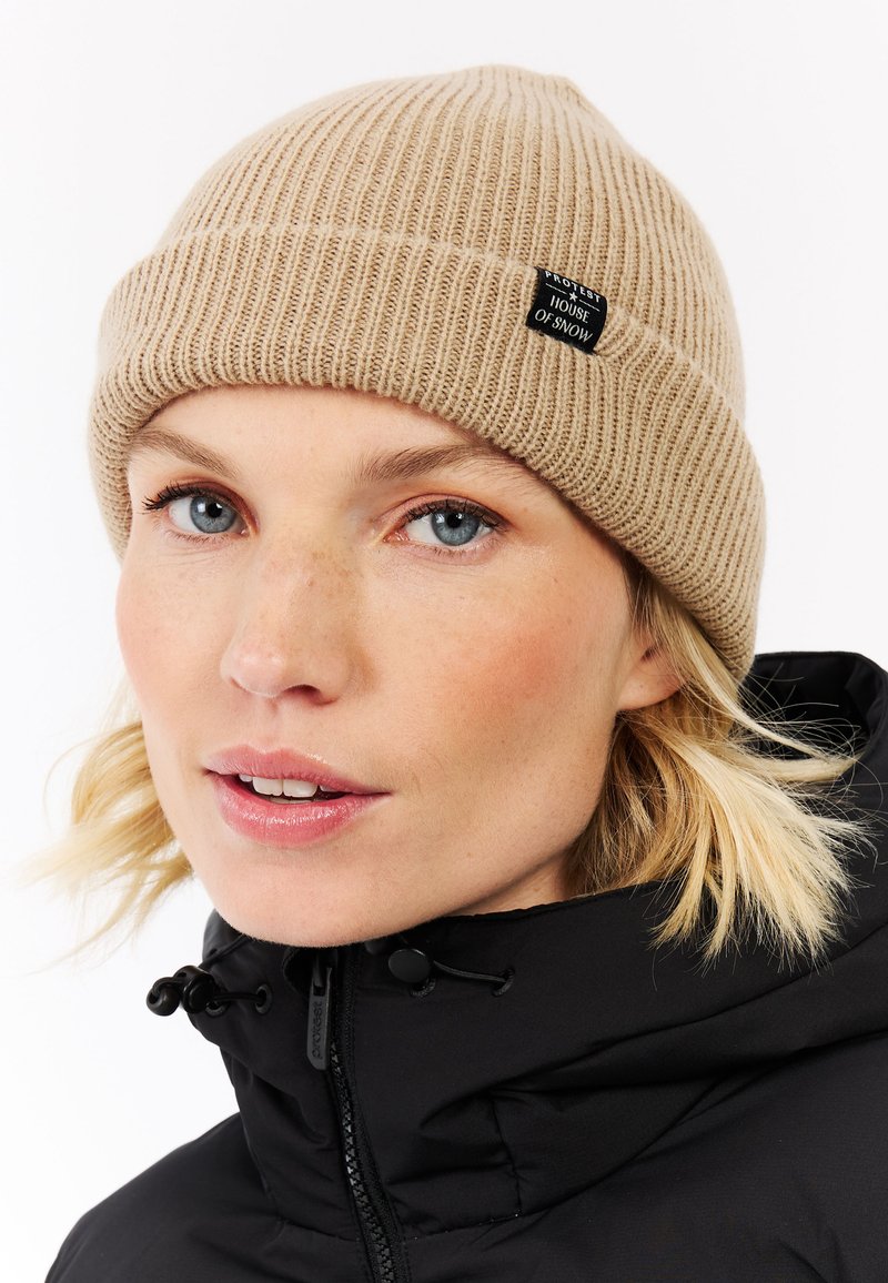 Tan ribbed beanie van gebreid materiaal, voorzien van een zwart label met witte tekst. Draagt bij een zwarte jas en toont een strakke pasvorm.