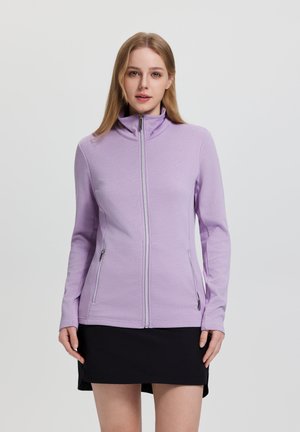 Junge Frau steht, trägt eine hellviolette Jacke mit Reißverschluss und zwei Reißverschlusstaschen sowie einen schwarzen Rock vor einem einfarbigen Hintergrund.