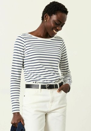 Gestreiftes Langarmshirt in Weiß und Marineblau mit rundem Ausschnitt. Kombiniert mit weißen Jeans und einem schwarzen Gürtel, der mit einer Hand gehalten wird.