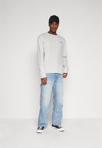 Lys grå langermet skjorte med blå logo, kombinert med løse lyseblå jeans og svarte joggesko. Capsen er vendt baklengs.