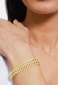 Złota bransoletka łańcuszkowa z ogniwami połączonymi ze sobą, o gładkiej, błyszczącej powierzchni, umieszczona na nadgarstku w kontekście teksturowanej skóry.