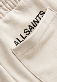 Tissu beige à texture lisse, avec une poche plaquée. Marque "ALLSAINTS" imprimée en noir sur la poche. Taille élastique en haut.
