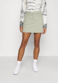 Donna che indossa una mini gonna di denim verde chiaro con tasche frontali, sneakers bianche alte e una maglietta a maniche lunghe tie-dye.
