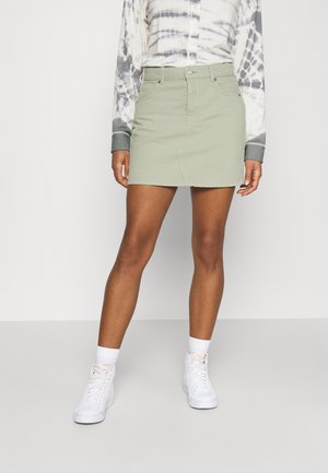 Donna che indossa una mini gonna di denim verde chiaro con tasche frontali, sneakers bianche alte e una maglietta a maniche lunghe tie-dye.