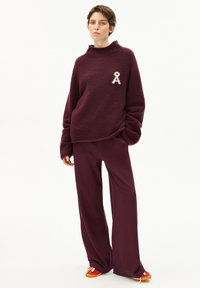 Burgunderfarbener Strickpullover mit hohem Kragen und weißen gestickten Bändern, kombiniert mit passenden burgunderfarbenen weit geschnittenen Hosen und mehrfarbigen Schuhen.
