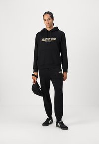 Fekete kapucnis pulóver és jogging szett, elülső zsebbekkel, sárga logóval és rugalmas mandzsettákkal. A modell fekete sapkát tart és fekete sportcipőt visel.