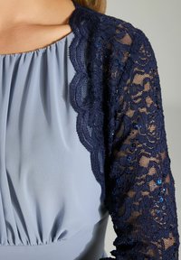Koronkowa kurtka z granatowym kwiatowym wzorem, cekinami i falbaniastymi brzegami na jasnoniebieskiej, marszczonej części górnej. Gładka tekstura materiału.