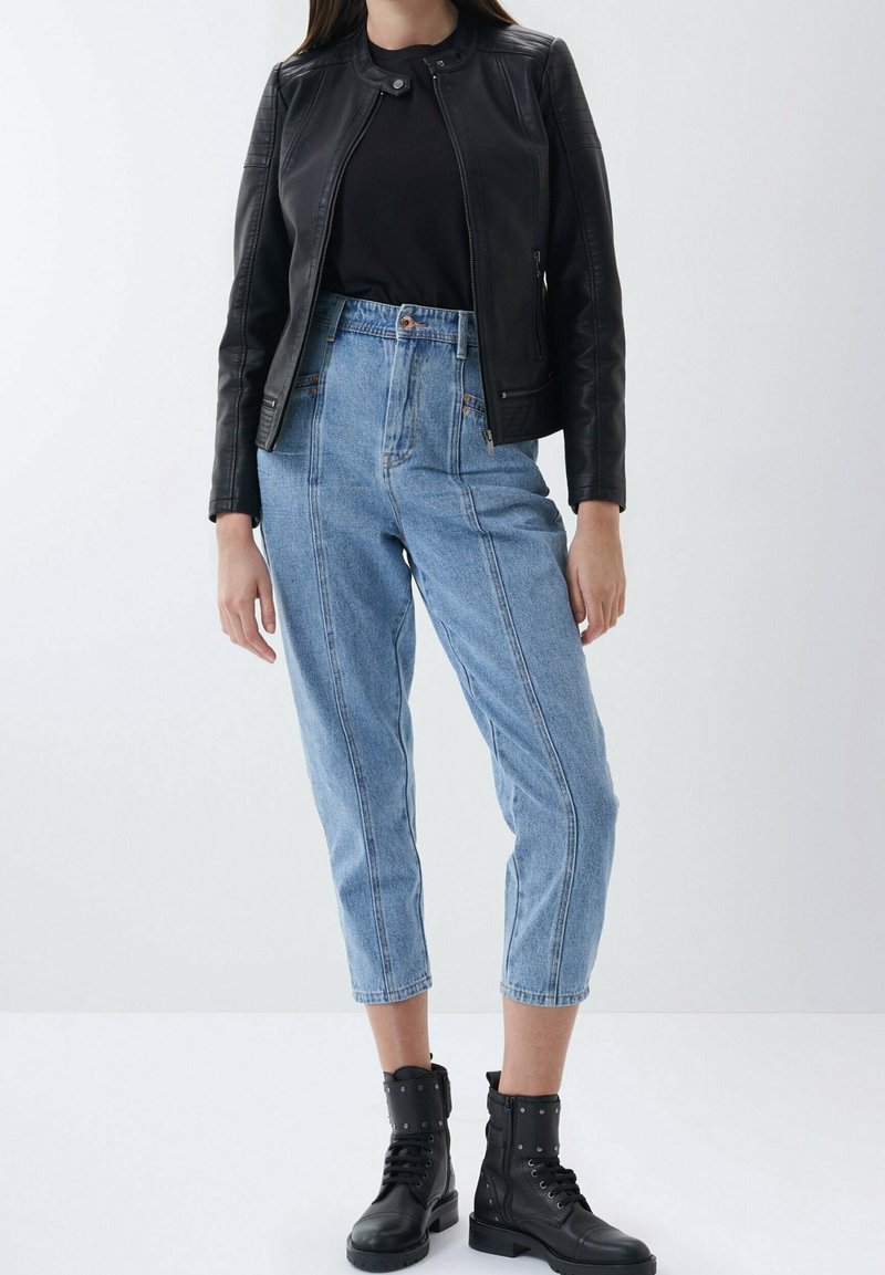 Donna con una giacca di pelle nera, top nero, jeans blu a vita alta e cropped, e stivali da combattimento neri con borchie, in piedi su un pavimento bianco.