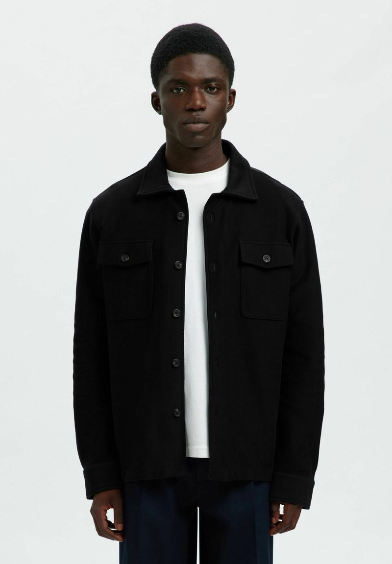 Noor mees kannab musta nööpidega jakki, millel on klapikotid, valge särgi peal, seistes lihtsa valge tausta ees.