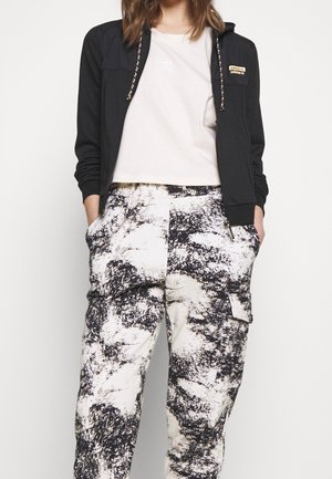 Femme portant un t-shirt court blanc, un sweat à capuche zippé noir avec logo et un pantalon cargo taille haute avec motif abstrait noir et blanc.