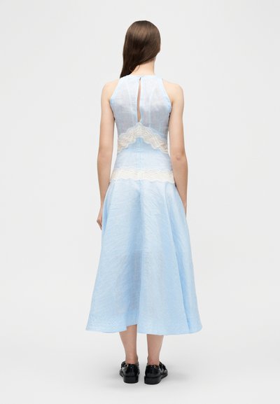 Γυναίκα που φοράει αμάνικο γαλάζιο φόρεμα με λευκές δαντελένιες λεπτομέρειες και μαύρα παπούτσια, στέκεται με την πλάτη στραμμένη σε απλό φόντο.