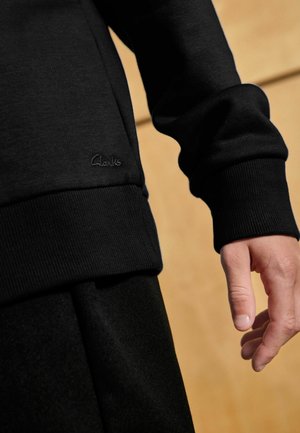 Sweatshirt noir avec des poignets côtelés et un petit logo "Clarks" brodé. Le matériau semble lisse et doux sur un fond texturé.