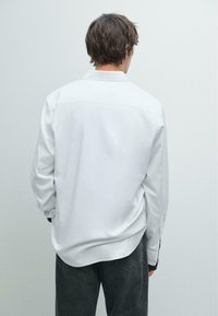 Weißes Langarmhemd mit lässiger Passform, mit Knopfmanschetten und geradem Saum, kombiniert mit dunkelgrauen Jeans. Glatte Stofftextur.