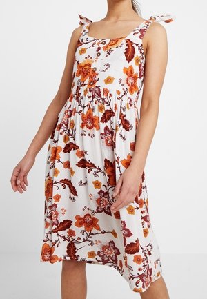 Biała sukienka na lato bez rękawów z pomarańczowo-brązowym kwiatowym wzorem, z przodu zapinana na guziki, z wiązanymi ramiączkami na stojącej kobiecie.