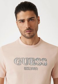 Camiseta de algodón rosa claro con un logotipo en relieve gris que dice "GUESS EST. 1981." Diseño de cuello redondo y mangas cortas. Textura suave.