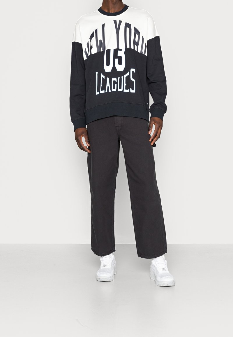 Sudadera en bloques de color blanco y negro con gráfico "NEW YORK 03 LEAGUES", combinada con pantalones negros de pierna ancha y zapatillas blancas.