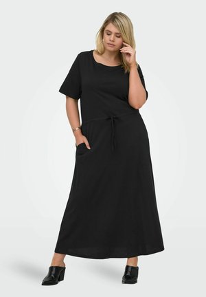 Donna con capelli biondi che indossa un vestito lungo nero a maniche corte e scarpe nere con il tacco, in piedi con una mano in tasca e l'altra che tocca il viso.