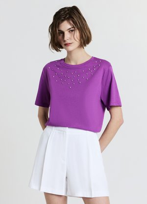T-shirt violet à manches courtes présentant un tissu texturé et un ensemble décoratif de clous en argent le long de l'encolure. Associé à un short blanc.