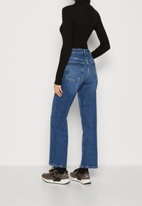 Žena v modrých vysokých straight-leg džínách, černém dlouhém roláku a tmavých teniskách, stojící na bílém pozadí.