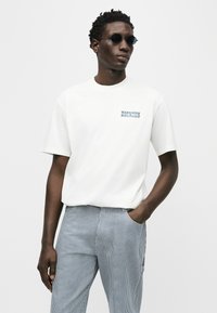 Άνδρας με σκούρα στρογγυλά γυαλιά ηλίου, λευκή κοντομάνικη μπλούζα Dickies και μπλε-άσπρο ριγέ παντελόνι, στέκεται με το ένα χέρι στην τσέπη.