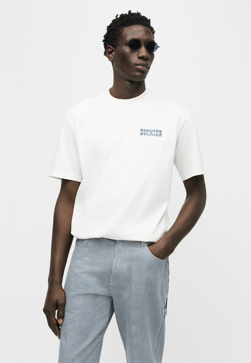 Man wearing dark round sunglasses, white short-sleeve Dickies t-shirt, and blue-and-white striped pants, standing with one hand in pocket.