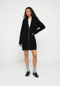 Μαύρη πλεκτή ζακέτα με φερμουάρ μπροστά, ριμπ υφή, μακριά μανίκια και χρυσές λεπτομέρειες στο φερμουάρ. Συνδυάζεται με γκρι κάλτσες και μαύρα μοκασίνια.