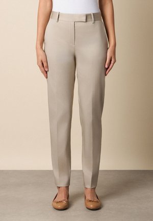 Femme portant un pantalon beige ajusté avec passants pour ceinture, pli devant et ballerines marron clair, debout devant un fond neutre.