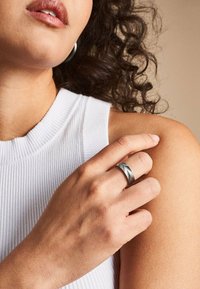 Silberner Ring mit einem Chevron-Muster, getragen an einer Hand, die auf einer nackten Schulter ruht. Das Model trägt ein weißes, geripptes, ärmelloses Oberteil.
