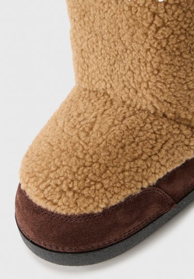 Botte en fausse fourrure beige avec une tige texturée, des accents en daim marron et une semelle extérieure en caoutchouc noir. Le design présente un bout rond et des détails de couture.