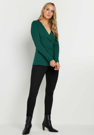 Green long-sleeve top with a deep V-neck and gathered detailing at the waist, paired with black skinny jeans and ankle boots.