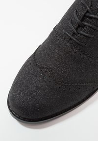Gros plan d'une chaussure oxford en suède noir avec une finition pailletée, des détails brogue et des lacets noirs sur fond blanc.