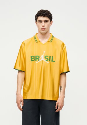 Ung mand med kort krøllet hår iført en gul Brasilien sportstrøje med grøn kant og hvid Jumpman-logo, stående mod en ensfarvet baggrund.